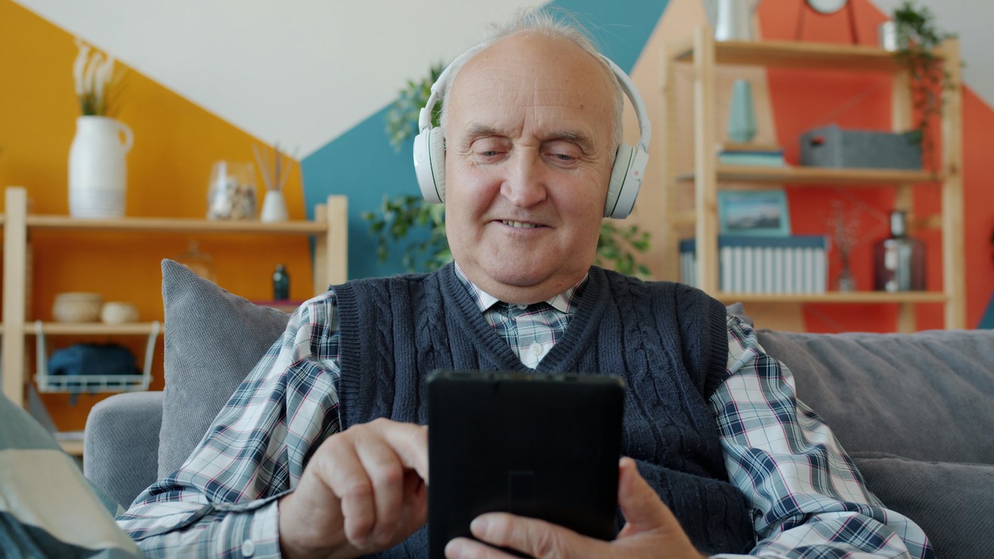 Starší muž se sluchátky na gauči se usmívá při používání tabletu.